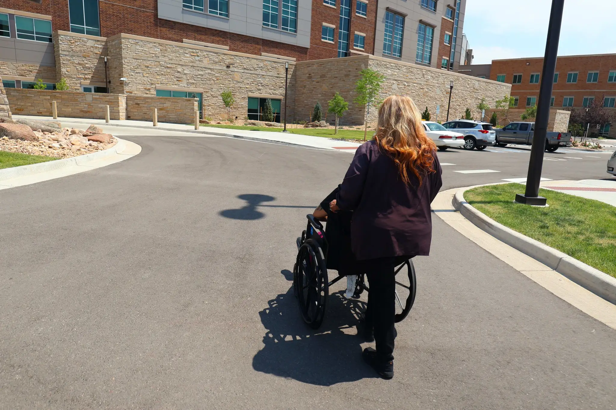 An attendant walking a passenger in a wheelchair along the pavement toward a hospital entrance.
